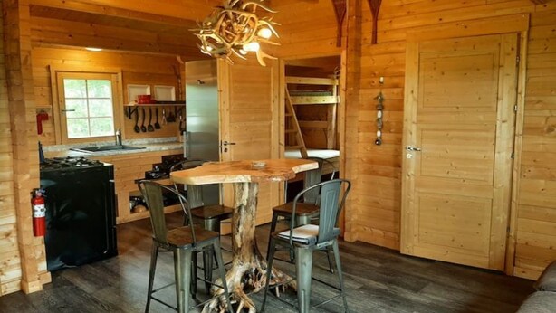 Un intérieur rustique en bois, avec une table à manger entourée de chaises, un lustre et un coin cuisine visible à travers une porte ouverte.
