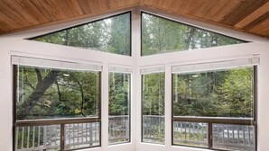 Una habitación de esquina con amplios ventanales, techo de madera y vista a árboles y una valla.
