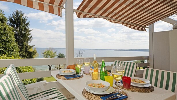 A terrace with striped outdoor furniture, a table set with plates of food, glasses of wine, and bottles. - Il Melagrano A11 by Wonderful Italy (Padenghe sul Garda)