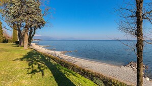 A lakeside view with a grassy area, trees, and a pebble beach. - Il Melagrano A11 by Wonderful Italy (Padenghe sul Garda)