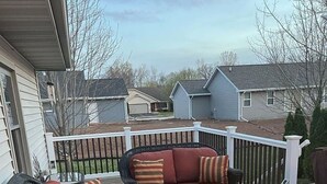 A deck with wicker furniture, including a sofa and chairs, and a glass-top table with a fire feature.