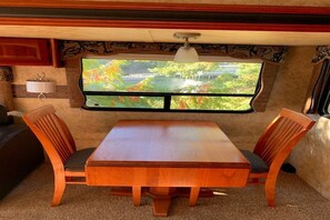 A compact RV interior with a dining area featuring a wooden table and chairs, a mounted TV displaying a scenic view, and a ceiling-mounted light fixture.