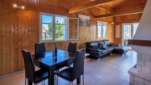 A spacious living area with a wooden ceiling, a dining table with chairs, a black leather sofa, and a fireplace.