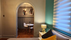 Une chambre d’hôtel comprenant un lit, une table de chevet avec une lampe, une fenêtre avec des stores, une petite table ronde avec des chaises, et donnant sur le couloir.