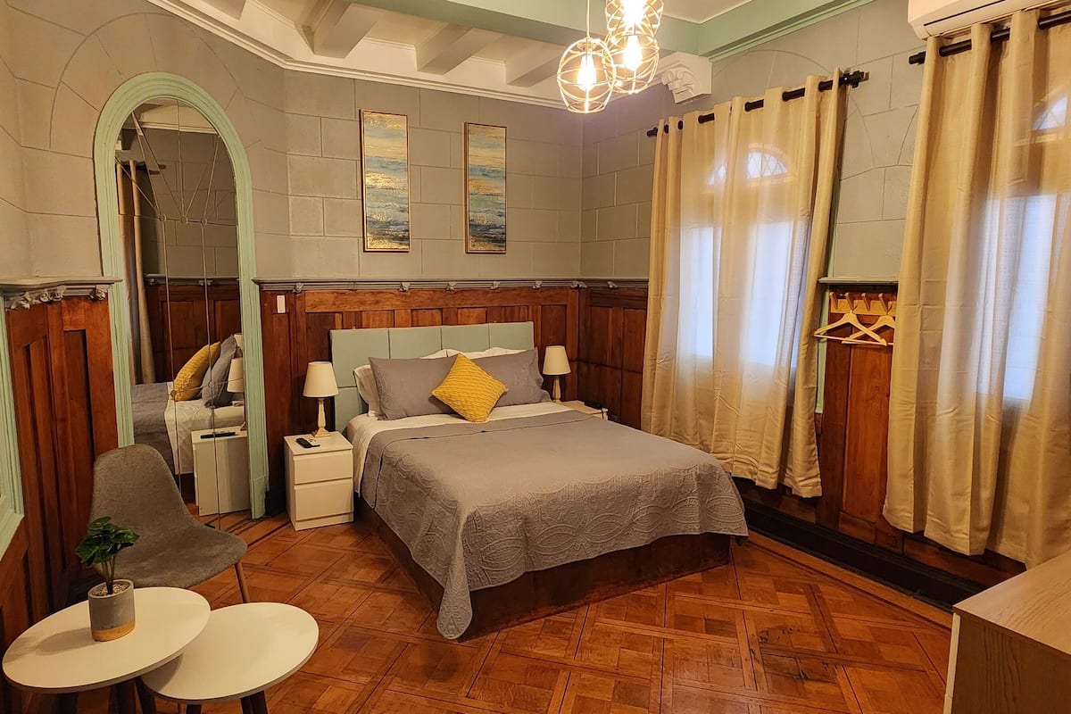 Ein Schlafzimmer mit einem großen Bett, einem Nachttisch, einem Sessel, einem kleinen Tisch mit einer Pflanze und einem Spiegel.