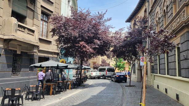 Une rue pavée bordée de cafés en plein air, de voitures garées et de bâtiments historiques.