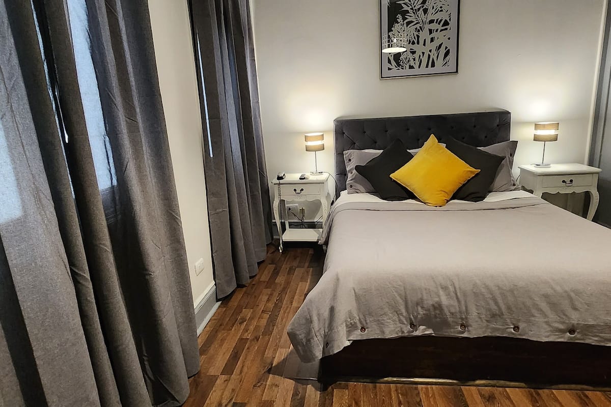 Ein Schlafzimmer mit einem großen Bett, grauen Vorhängen, einem Nachttisch mit Lampe sowie einem an der Wand hängenden Kunstwerk.