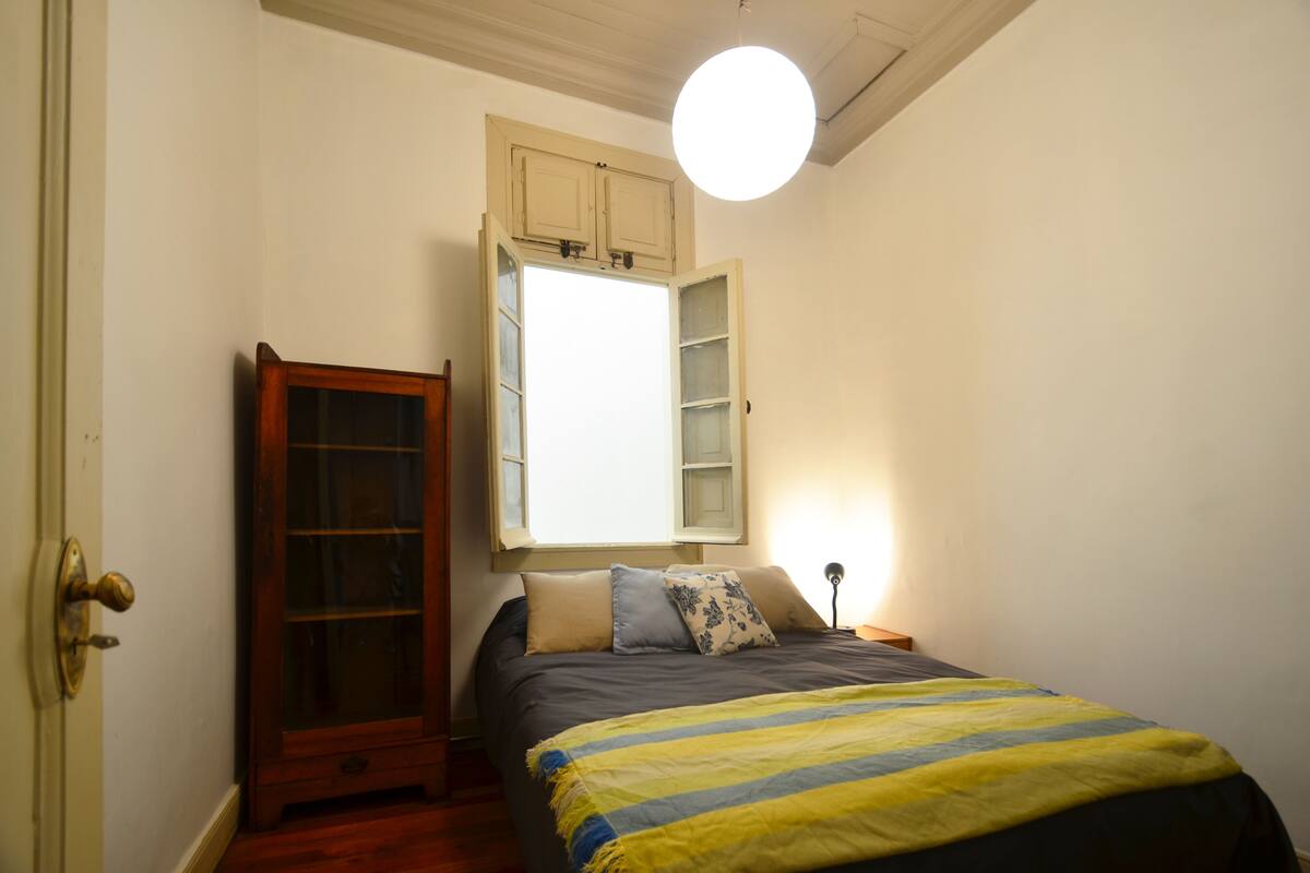 A bedroom with a bed, a wooden cabinet, a window with open shutters, and a round ceiling light.