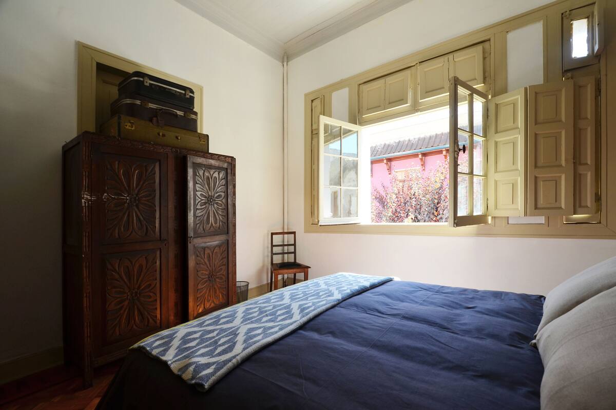 A bedroom with a bed, a wooden wardrobe, a chair, and a window with open shutters.