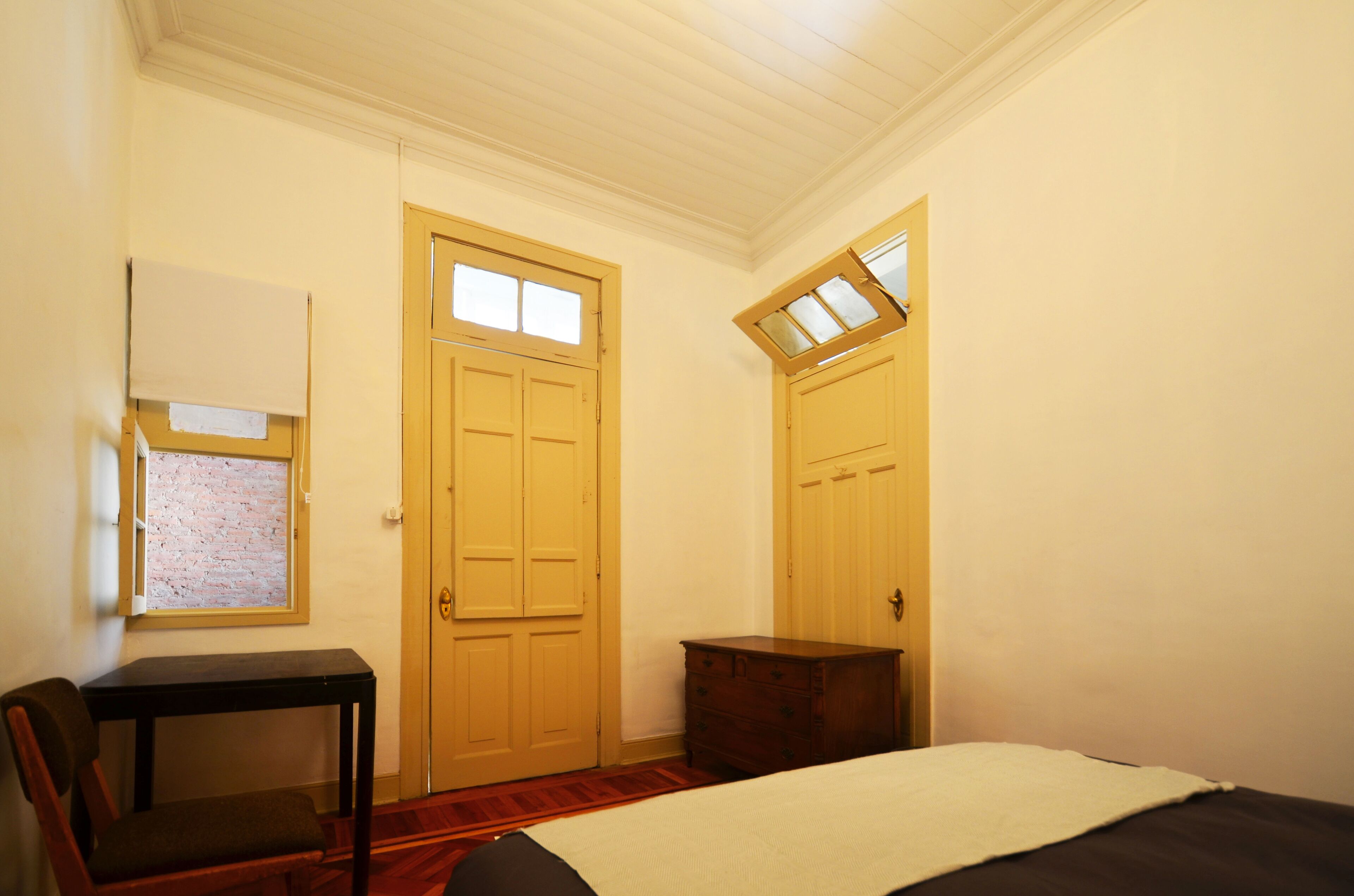Een slaapkamer met een bed, een houten dressoir, een stoel, een bureau en twee deuren met ramen.