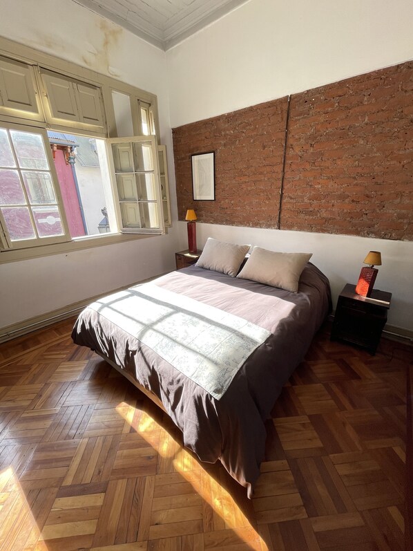 A bedroom with a herringbone wooden floor, a bed with a patterned bedspread, a brick accent wall, and a window with a view of the outside. - CASA LUCRECIA (Santiago)