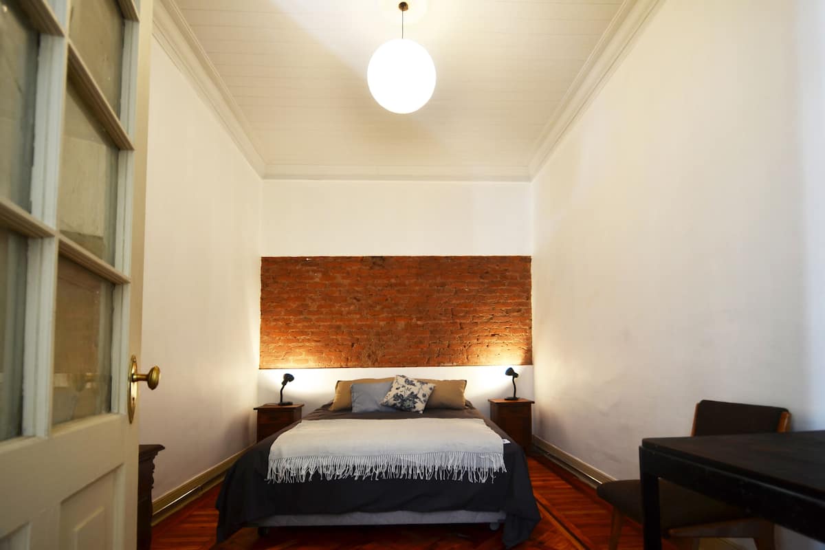A bedroom with a brick accent wall, a bed with a white and black bedspread, two bedside lamps, and a wooden floor.