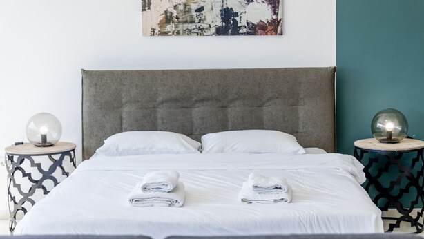 A neatly made bed with two folded towels, a grey upholstered headboard, and two bedside tables with decorative glass lamps. - Hip And Fun in Athens Apt B (Athens)