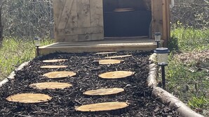 Eine Holzhütte mit einer Tür, umgeben von einem Gartenpfad mit runden Trittsteinen.