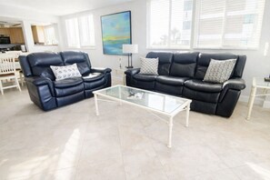 A modern living room with two black leather sofas, a glass coffee table, and a dining area in the background. - Spacious Beachfront Corner Condo in Madeira Beach| Gulf-Front|Pool & Hot-tub (Madeira Beach)