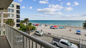 A beach view from a balcony with a white building, palm trees, and beach umbrellas. - Spacious Beachfront Corner Condo in Madeira Beach| Gulf-Front|Pool & Hot-tub (Madeira Beach)