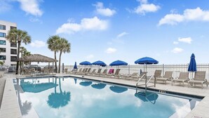 A swimming pool with lounge chairs and umbrellas, surrounded by palm trees and a white fence. - Spacious Beachfront Corner Condo in Madeira Beach| Gulf-Front|Pool & Hot-tub (Madeira Beach)