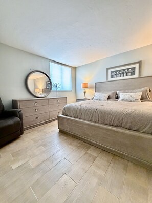 A bedroom with a bed, dresser, chair, and a window with blinds. - Spacious Beachfront Corner Condo in Madeira Beach| Gulf-Front|Pool & Hot-tub (Madeira Beach)