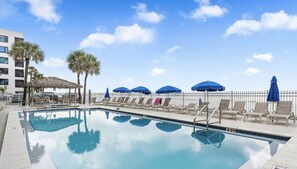 A swimming pool with lounge chairs and umbrellas, surrounded by palm trees and a white fence. - Spacious Beachfront Corner Condo in Madeira Beach| Gulf-Front|Pool & Hot-tub (Madeira Beach)