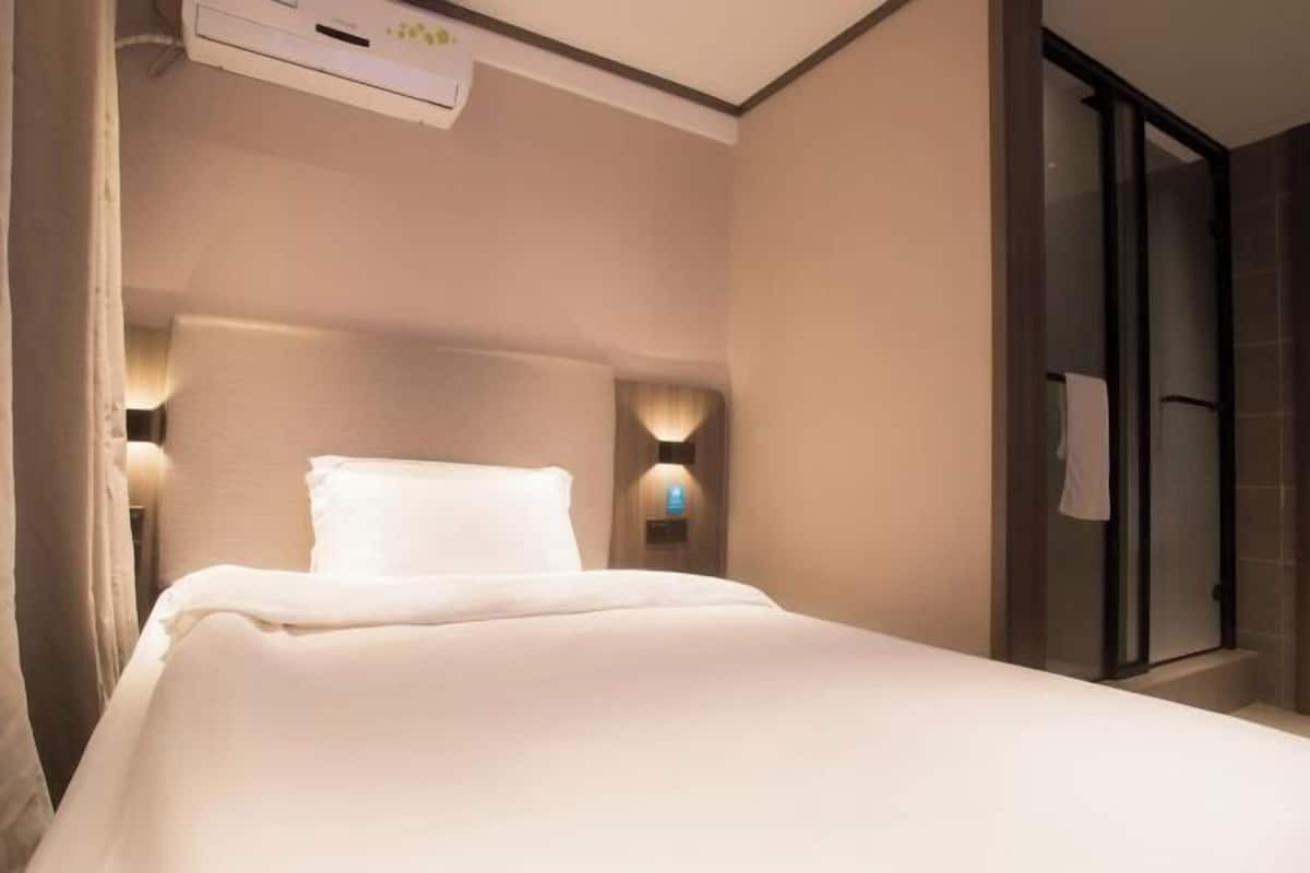 A hotel room with a bed, a wall-mounted air conditioner, and a glass shower enclosure.