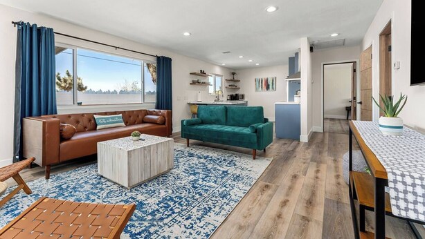 A modern living room with a brown leather sofa, teal sofa, and a coffee table. There is a kitchen area with a blue countertop and a dining table with a patterned table runner.