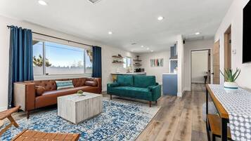 A modern living room with a brown leather sofa, teal sofa, and a coffee table. There is a kitchen area with a blue countertop and a dining table with a patterned table runner.