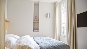 A neatly made bed with white linens and a gray blanket, a small shelf with a vase, and a window with curtains. - Amazing Apt Prestige at Mouffetard Paris 5 (Paris)