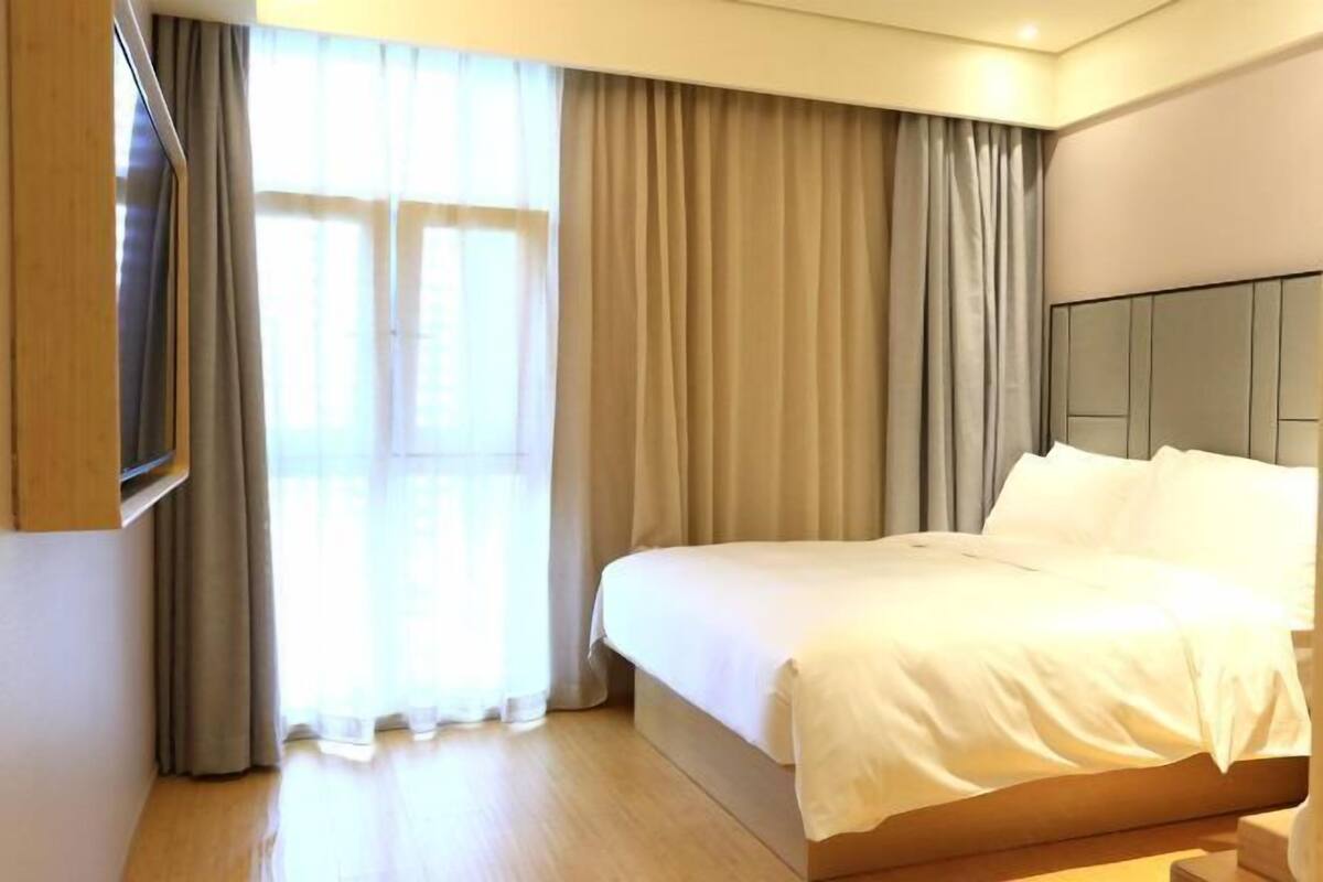 A hotel room with a large bed, a flat-screen TV mounted on the wall, and a large window with curtains.