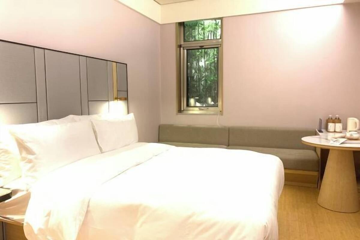 A hotel room with a large bed, a bench, a round table, and a window with a view of greenery.