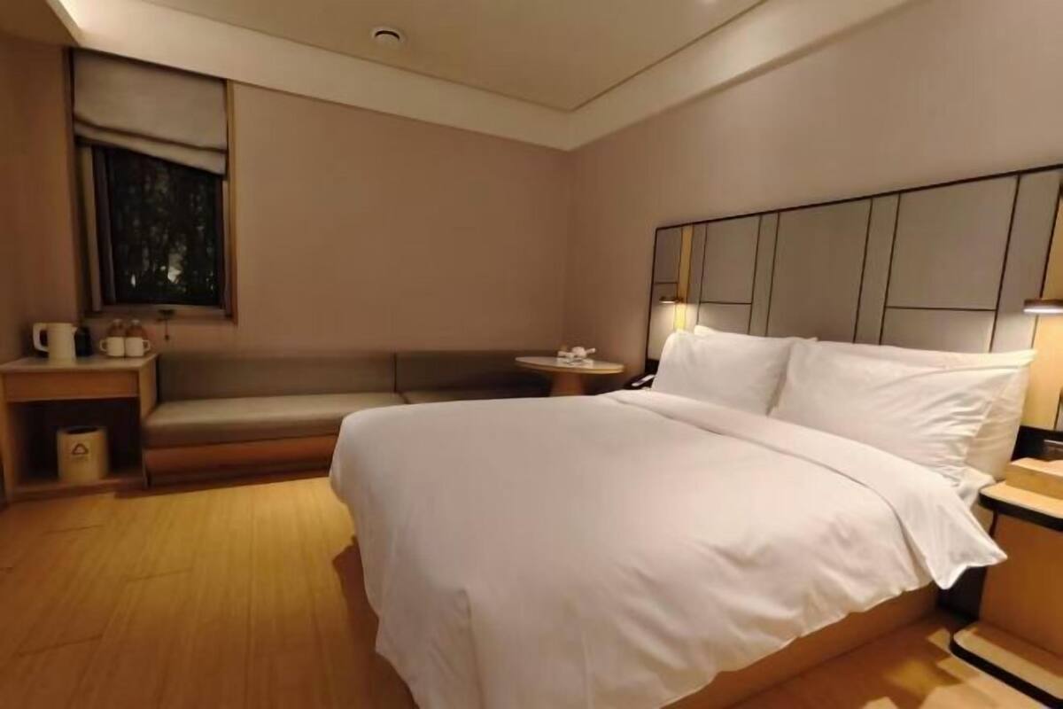 A hotel room with a large bed, a bedside table, a small table, a bench, and a wall-mounted mirror.