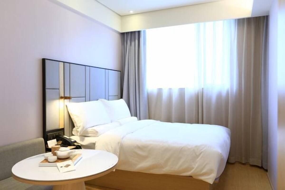 A hotel room with a bed, a bedside table with a lamp, a round table with books and a bowl, and a large window with curtains.