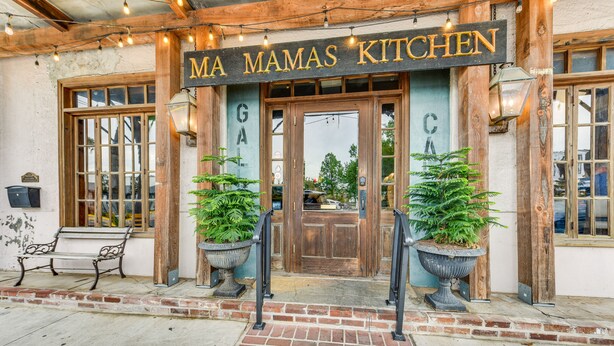 L'entrada de 'MA MAMAS KITCHEN' amb un camí de maons, portes de fusta i dues plantes grans en testos.