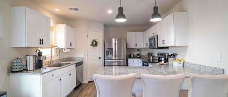 Una cocina moderna con armarios blancos, encimera de granito, electrodomésticos de acero inoxidable e iluminación colgante.