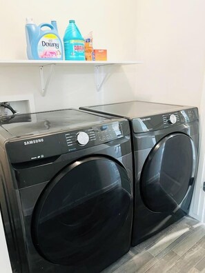 A Samsung front-loading washer and dryer set in a laundry room with a white shelf above containing cleaning products. - 4BR Casa Grande Home | Fast WiFi, Grill + Sleeps 8 (Casa Grande)
