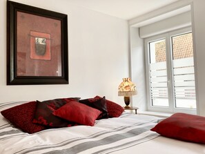 A bedroom with a bed, a framed picture on the wall, a lamp, and a window with blinds. - Le Familik - Maison - Centreville (Licques)