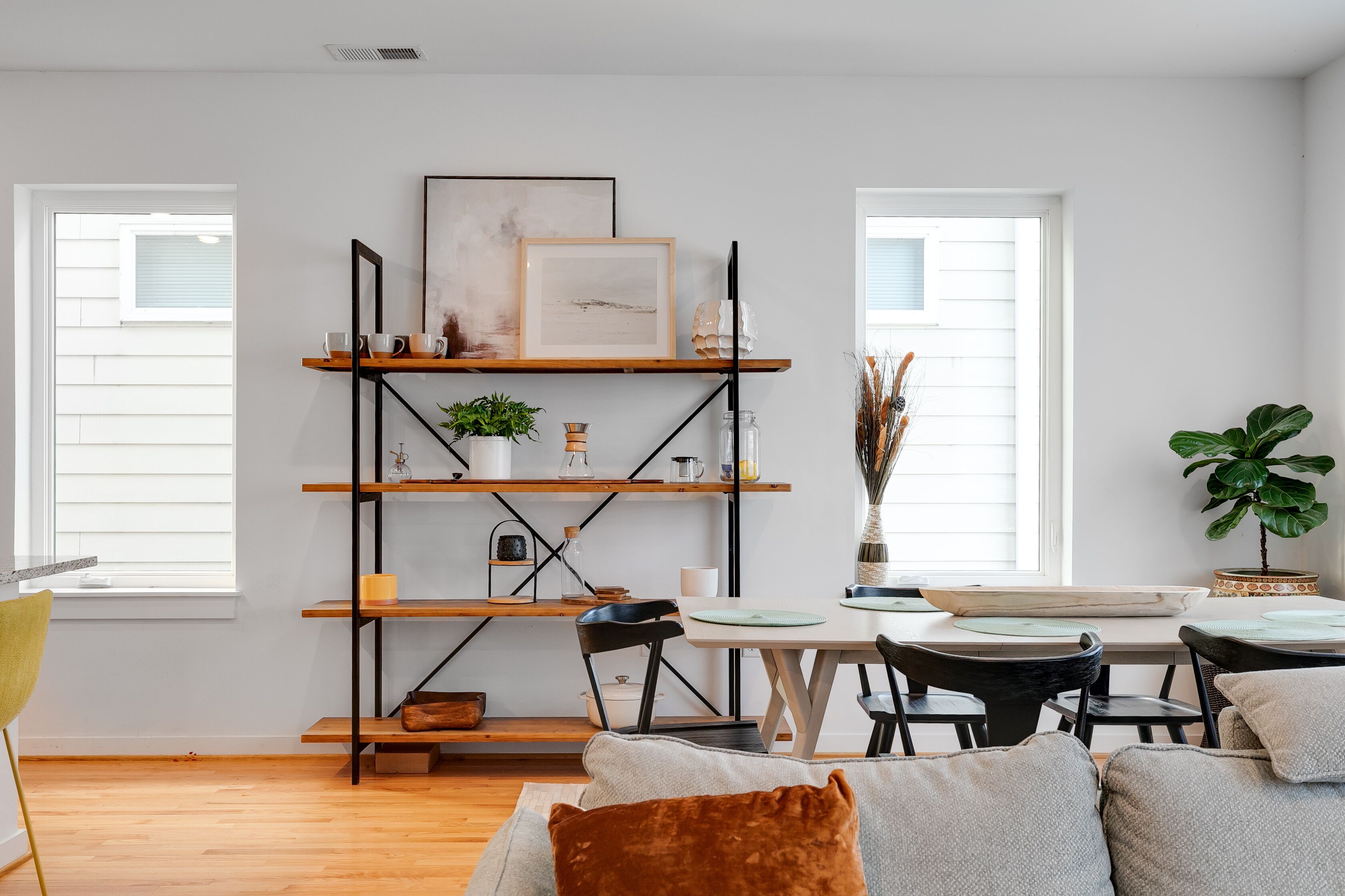 A modern living room with a wooden floor, a sofa with cushions, a dining table with chairs, a bookshelf with various items, and two windows with blinds.