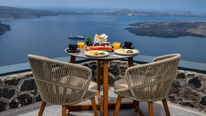 A table with two wicker chairs, plates of food, and glasses of orange juice, overlooking a scenic body of water. - Eteoro Suites (Santorini)