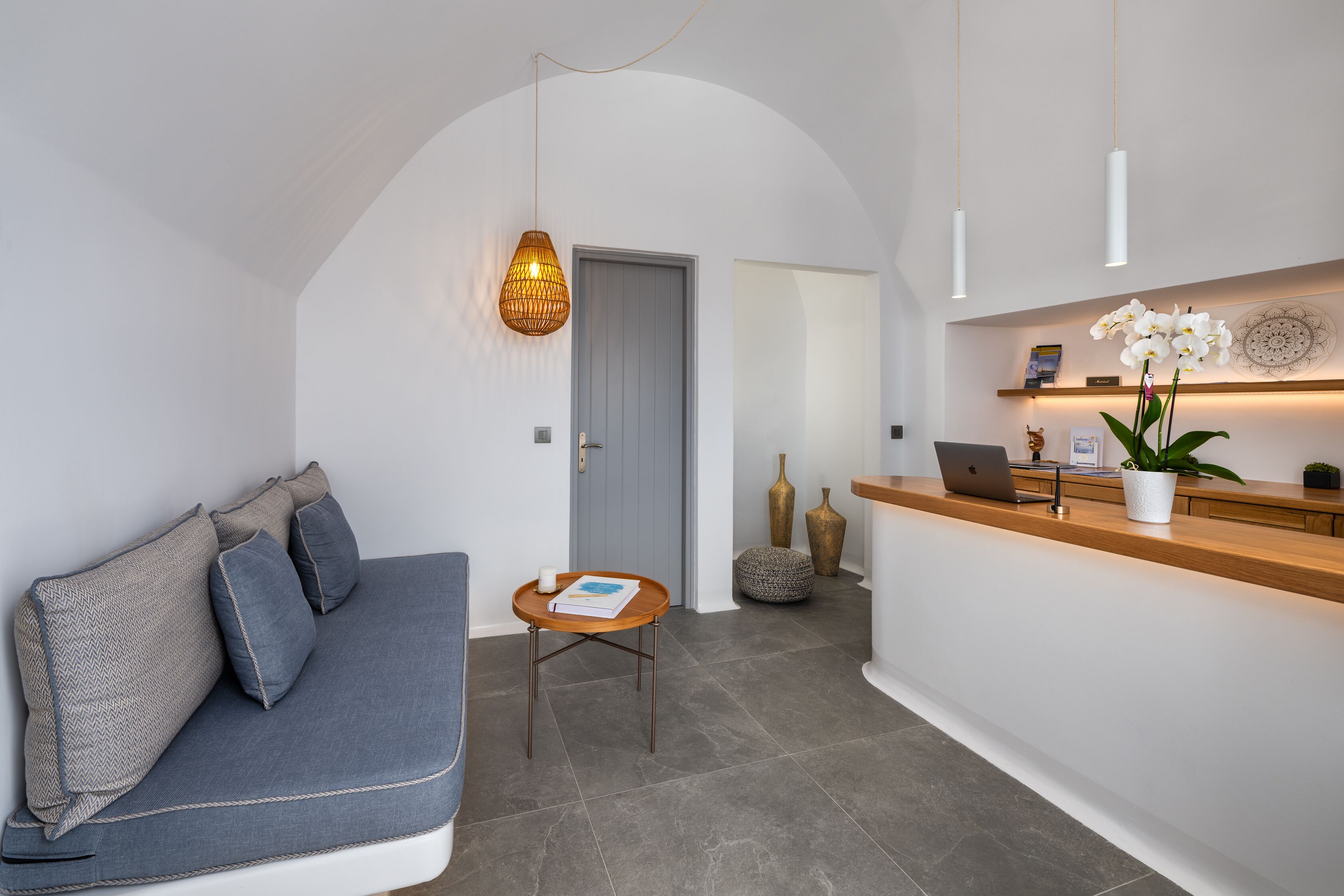 Un espace d’accueil moderne avec un comptoir de réception en bois, un canapé gris, une petite table ronde avec un livre et un luminaire suspendu.