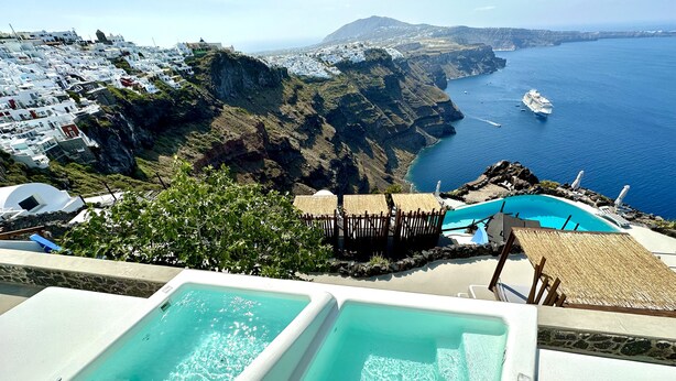 A pool area with clear blue water, overlooking a coastal landscape with white buildings and a view of the sea. - Eteoro Suites (Santorini)