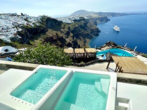 Zona de piscina con agua azul cristalina, con vistas a un paisaje costero de edificios blancos y al mar.
