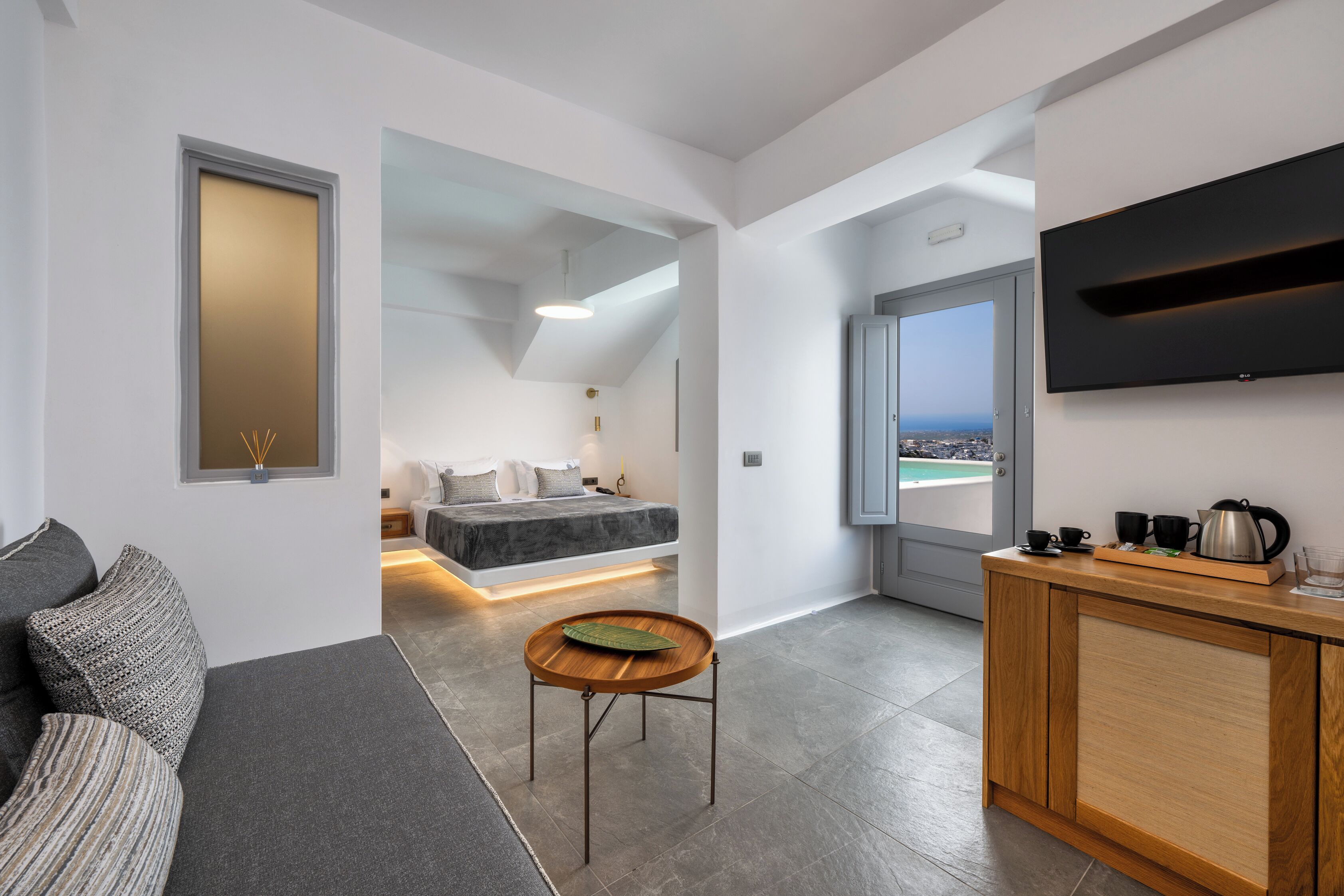 A modern, compact living space with a sofa, a small wooden table, a bed with a view, a kitchenette, and a TV mounted on the wall.
