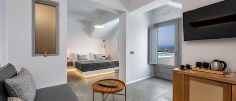 A modern, compact living space with a sofa, a small wooden table, a bed with a view, a kitchenette, and a TV mounted on the wall.