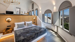 A modern bedroom with a large bed, a wooden headboard, a TV, and a view of the landscape. - Eteoro Suites (Santorini)