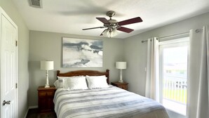 A bedroom with a bed, two bedside tables with lamps, a ceiling fan, a window with curtains, and a painting on the wall.