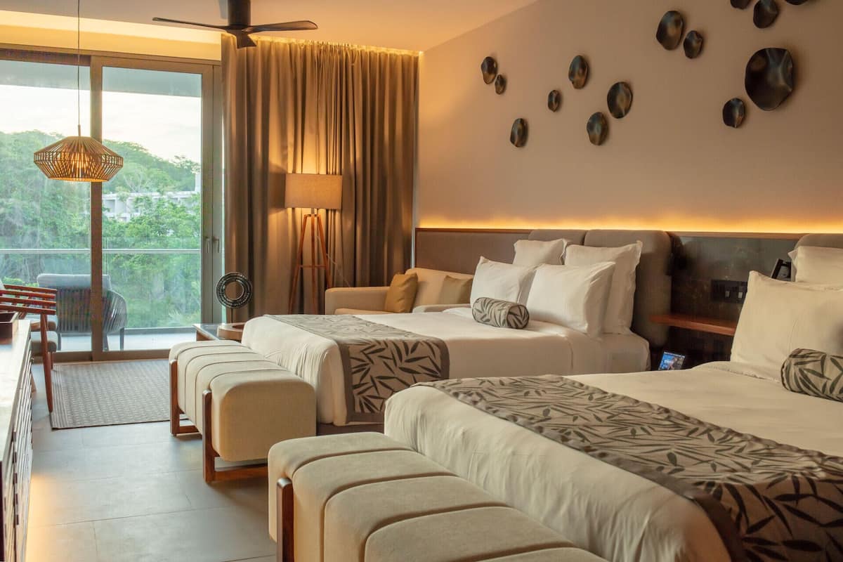 A hotel room with two beds, a large window with curtains, a ceiling fan, and a view of greenery outside.