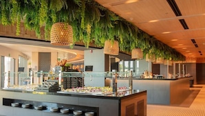 A buffet area with a variety of food items, a glass display case, and hanging greenery. - Secrets Bahia Mita Surf & Spa (Punta de Mita)