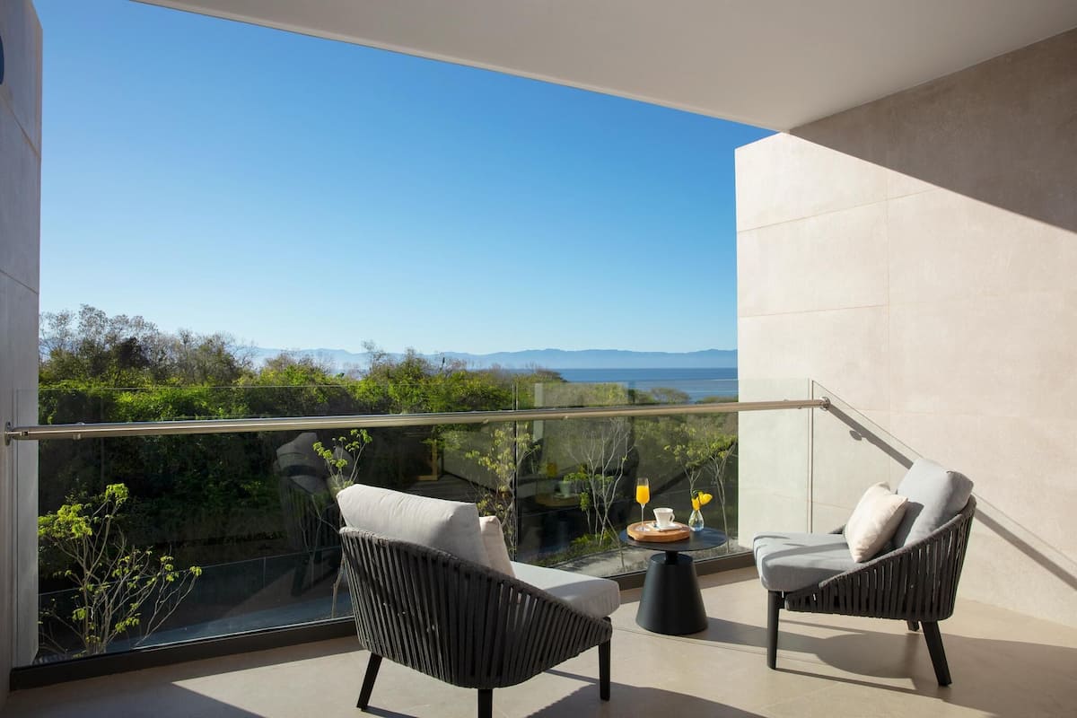 A balcony with two wicker chairs, a small round table with a tray, and a glass railing overlooking a lush landscape with distant mountains.