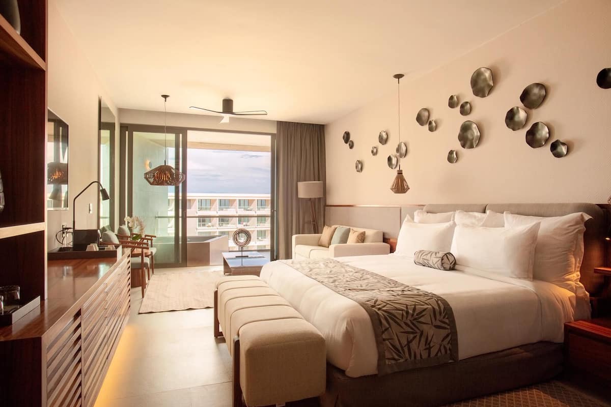 A modern hotel room with a large bed, a seating area with a sofa and chairs, a desk, and a view of the city through large windows.