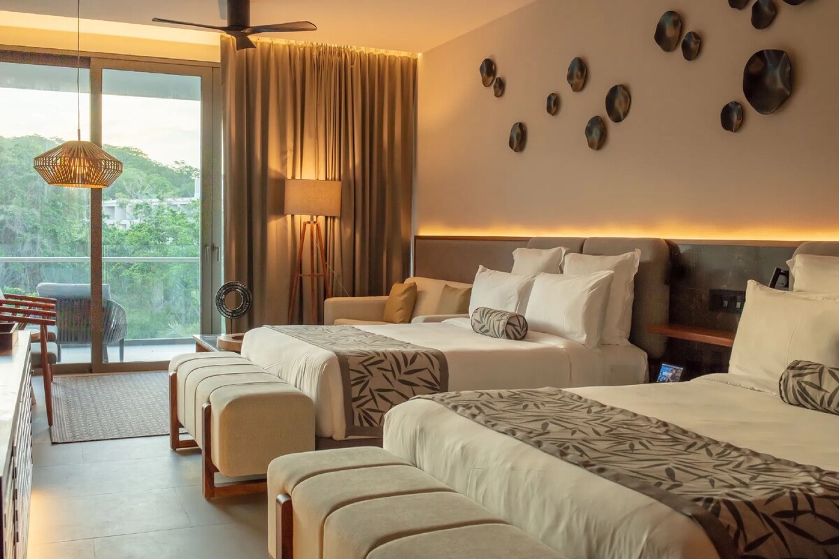 Quarto de hotel com duas camas, varanda, ventoinha no teto e vista para o verde.