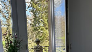 A room with a large window, a radiator, a bed with a grey quilt, and a vase with flowers on the windowsill.